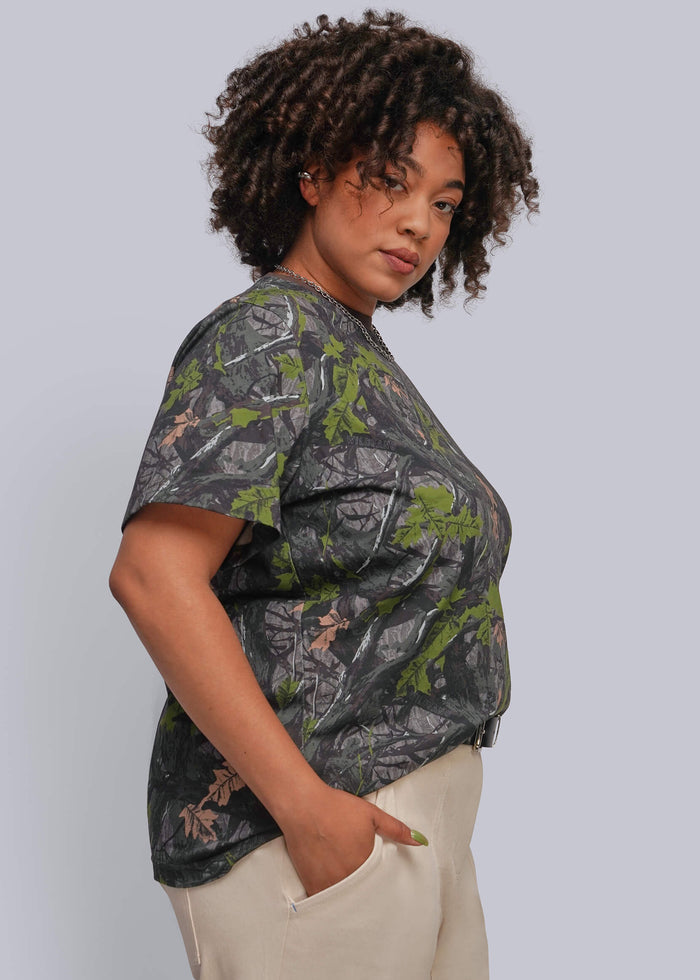 1X=A woman wearing a short-sleeve crewneck top with a dark gray and green camouflage pattern, paired with beige pants, photographed against a solid light gray background.