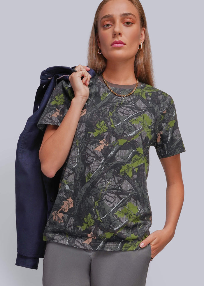 S=A woman modeling a short-sleeve crewneck t-shirt with a dark gray and green forest camouflage pattern, holding a navy blue jacket over her shoulder against a solid gray studio background.