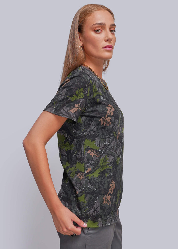 S=A woman modeling a short-sleeve V-neck top with a dark gray and green camouflage pattern, paired with gray pants, against a solid light gray background.