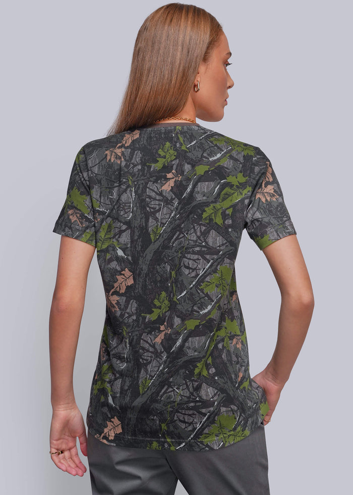 S=A woman shown from the back wearing a short-sleeve camouflage t-shirt with a realistic tree bark and leaf pattern in dark gray, green, and brown, paired with gray pants, against a solid gray studio background.