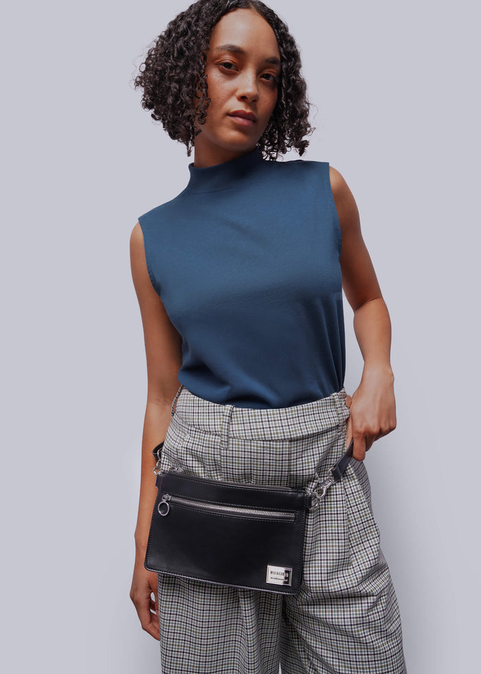 Woman wearing a blue sleeveless top and plaid pants with a black belt bag against a gray background