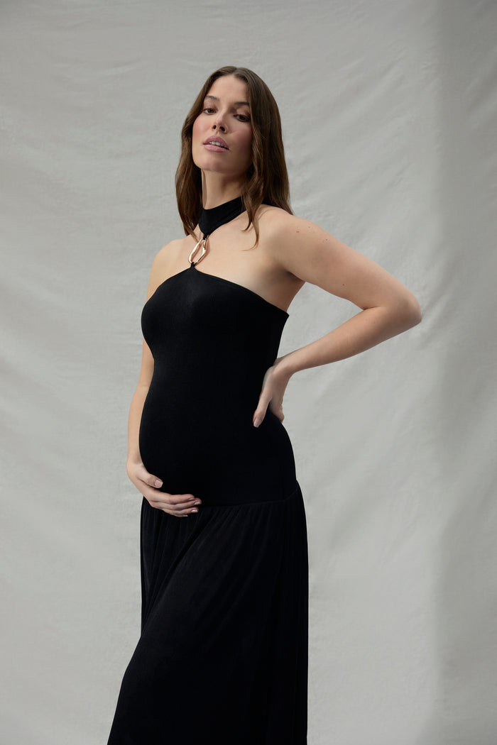 Pregnant woman wearing a black strapless dress with a gold halter accent against a plain background