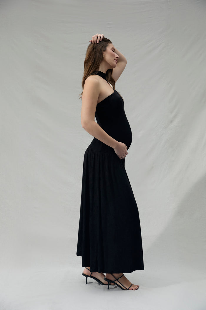 Pregnant woman wearing a black strapless hair dress with a gold halter accent against a plain background