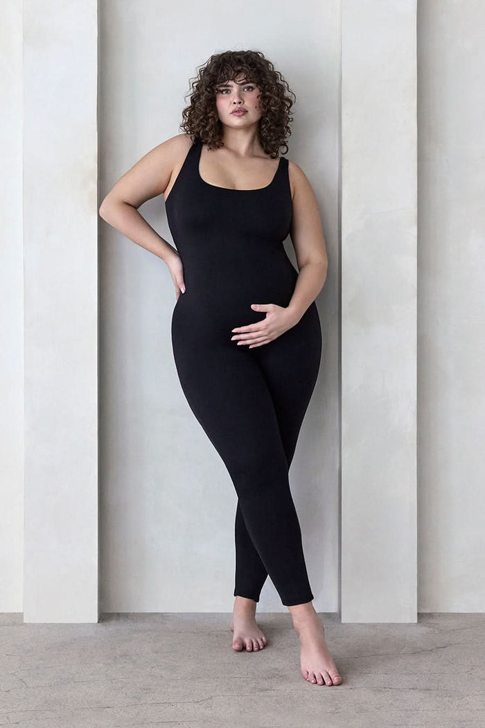 A curvy woman wearing the umpsuit Maternity Lucy Sleeveless Scoop Neck Jumpsuit in Black standing infront of a feature wall