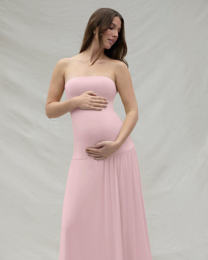 Pregnant woman wearing a pink strapless gown by Bumpsuit against a beige background