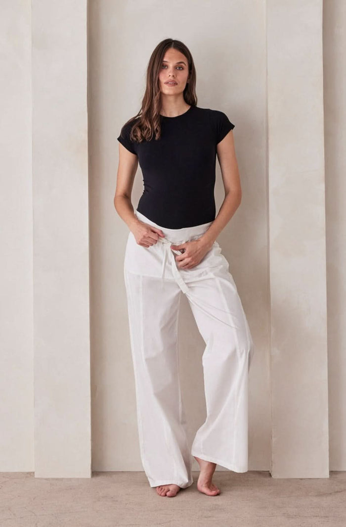 A pregnant woman wearing white relaxed pants and a black shirt by Bumpsuit against a beige feature wall.