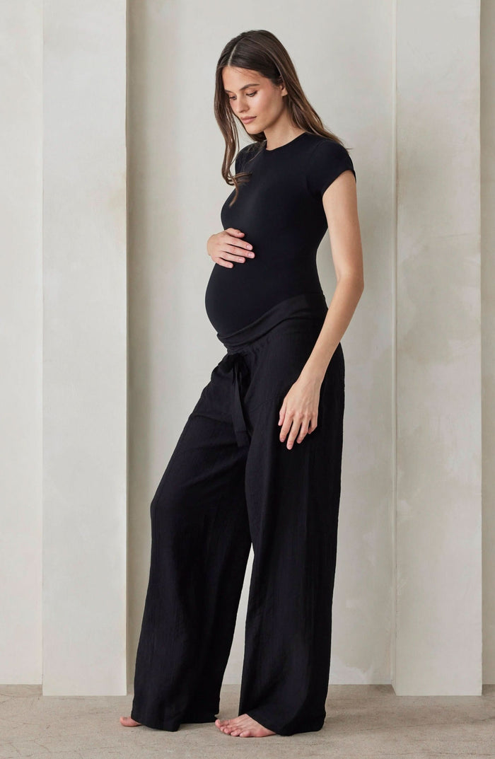 A pregnant woman wearing black relaxed pants and a black shirt by Bumpsuit against a beige feature wall.