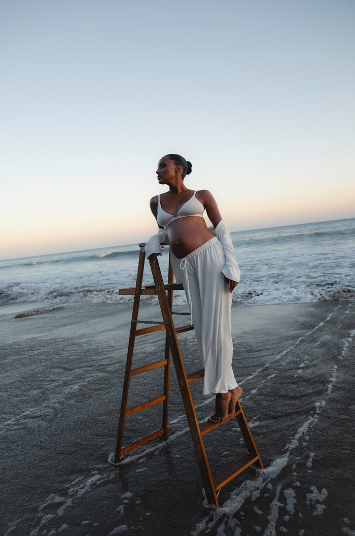 Pregnant woman standing on a beach with a wooden ladder, wearing white bumpsuit pjs and bra.