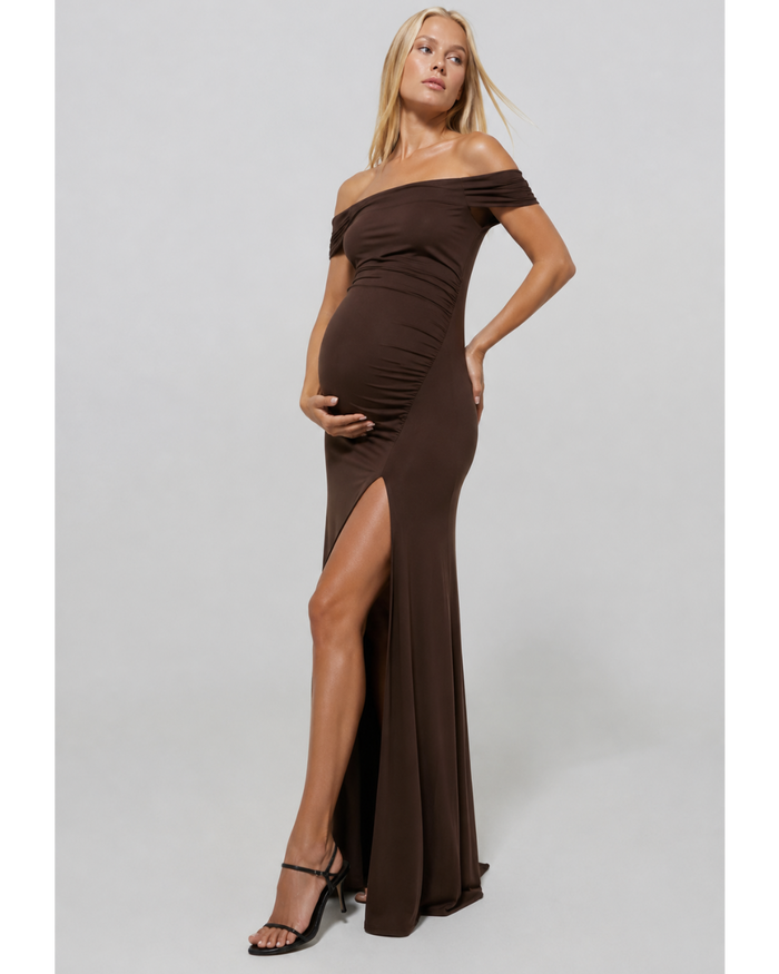 Pregnant woman wearing a chocolate coloured Bumspuit off the shoulder evening dress with a high leg split, set against a white background.