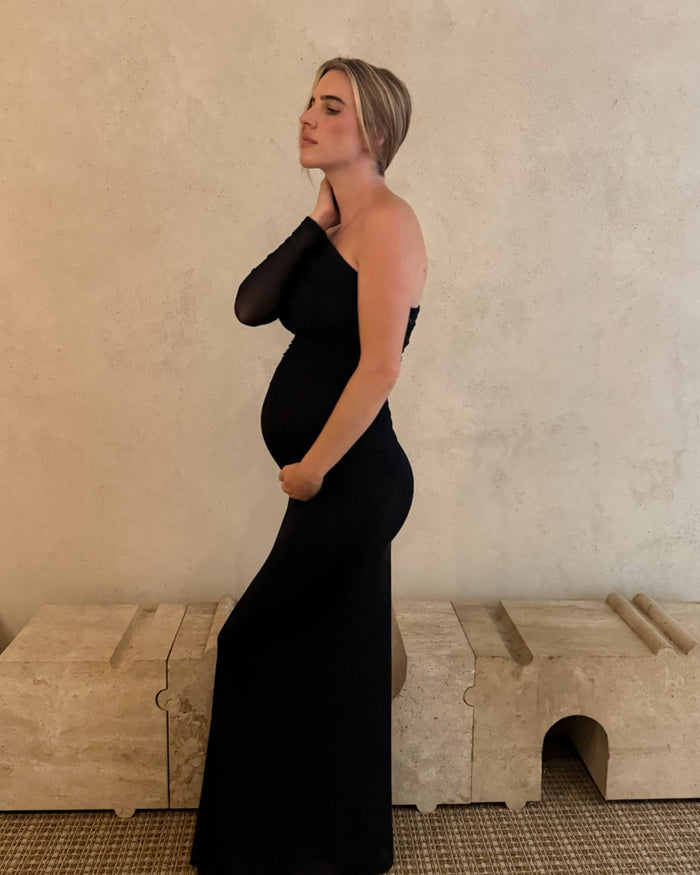 A pregnant woman wearing a black one-shoulder maternity gown by Bumpsuit stands in profile against a textured beige wall, with a light stone bench and woven rug in the background.
