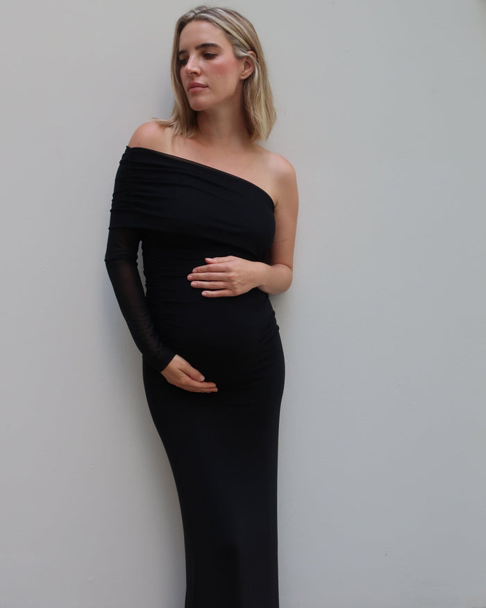 A woman wearing a Bumpsuit black off-the-shoulder long-sleeve maternity gown, standing against a plain light gray wall.