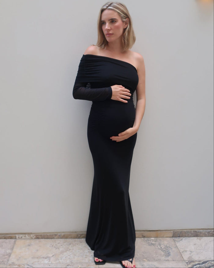 A pregnant woman wearing a black off-the-shoulder long-sleeve maternity gown by Bumpsuit, standing against a light gray wall.