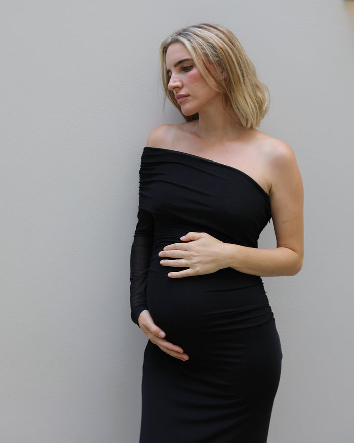 A pregnant woman with blonde hair wearing a black off-the-shoulder long-sleeve maternity gown by Bumpsuit, posing with her hands on her belly against a light gray wall.