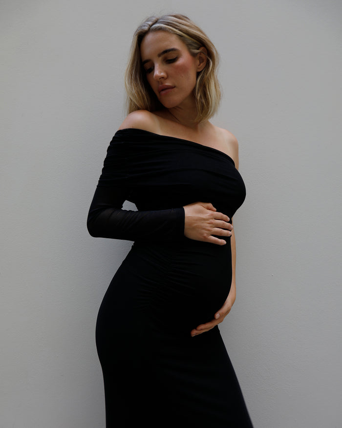 A pregnant woman wearing a Bumpsuit black off-the-shoulder long-sleeve maternity dress, posing with her hands on her belly against a plain gray wall.
