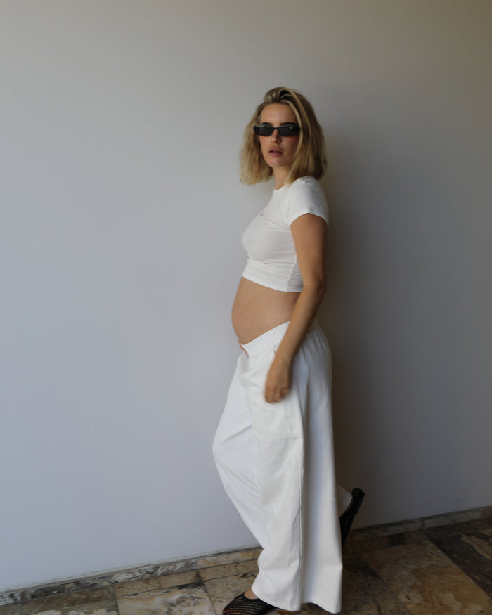 A pregnant woman with blonde hair and sunglasses stands in profile wearing a ivory short-sleeve crop top and matching white wide-leg pants  by Bumpsuit against a plain light gray wall.