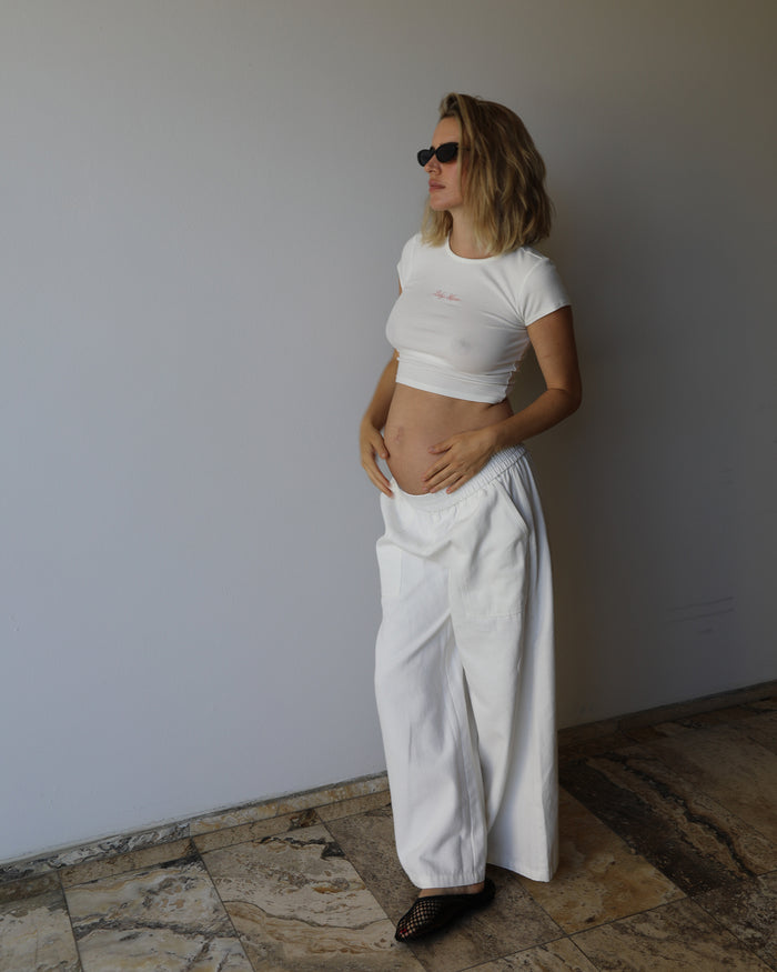 A woman wearing a Bumpsuit baby mama tee and matching white wide-leg jeans, standing in a minimalist room with a light gray wall and stone tile floor.