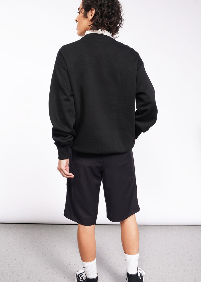 S=A person with curly hair stands against a plain background, dressed in a black Wild Feminist Fleece Crew, black shorts, white socks, and black sneakers. The brushed fleece adds comfort while subtly capturing the spirit of a WILD FEMINIST.