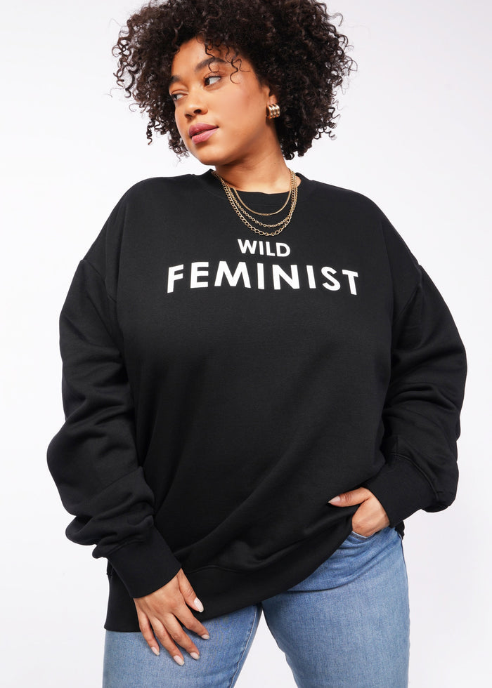 1X=Against a plain white backdrop, someone sports the Wild Feminist Fleece Crew in black. The chic look is accentuated by bold WILD FEMINIST text, their curly hair, gold hoop earrings, and layered necklaces. The outfit is elevated by the crews soft brushed fleece feel.