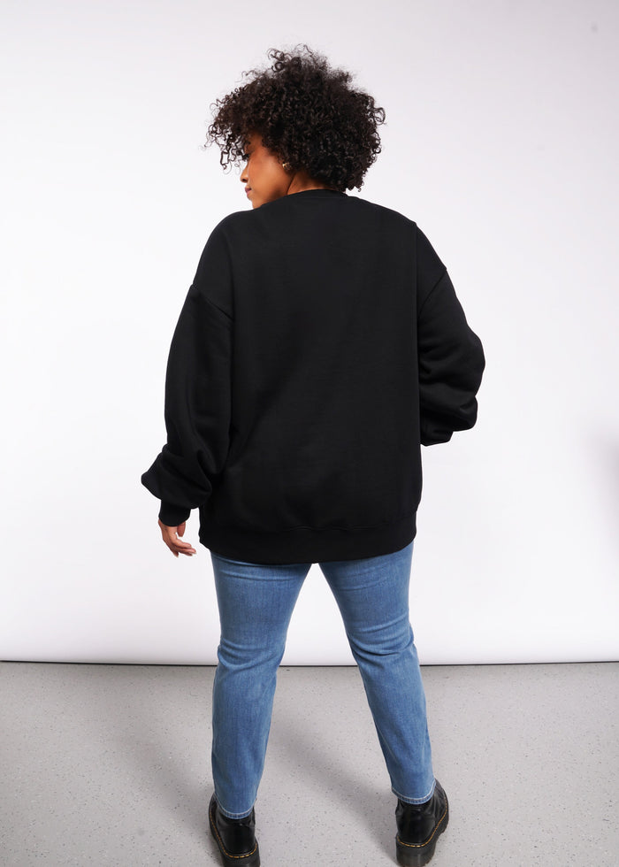 1X=In a white room, a person with curly hair faces away wearing the Wild Feminist Fleece Crew paired with blue jeans and sturdy black boots.