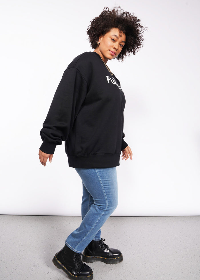 1X=A person casually leans against a white background, wearing the Wild Feminist Fleece Crew in black with white lettering, paired with blue jeans and black boots. The brushed fleece enhances the texture of their look. They have curly hair and gaze slightly to the side.