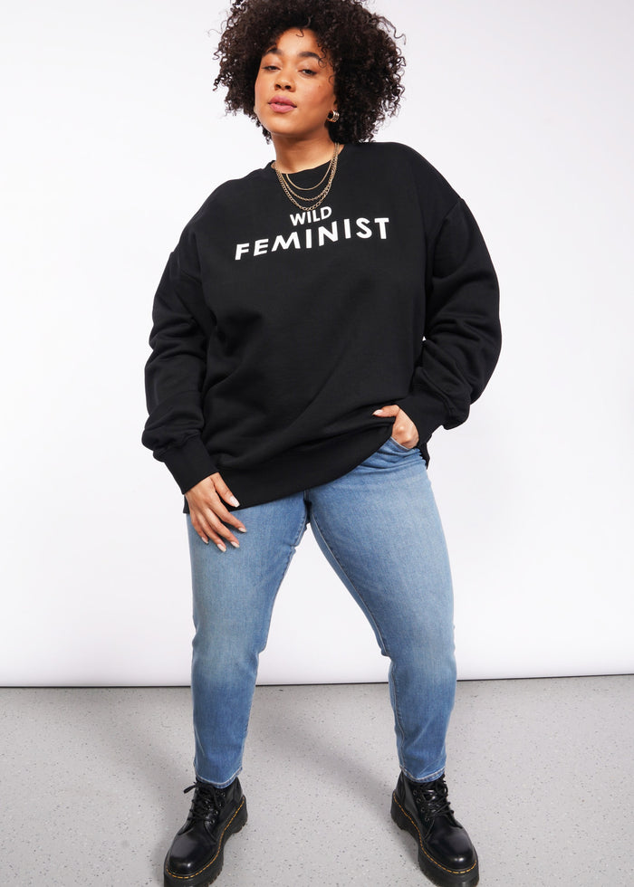 A person with curly hair confidently wears the Wild Feminist Fleece Crew paired with light blue jeans. Black boots complete their strong and stylish look as they rest a hand on their hip.