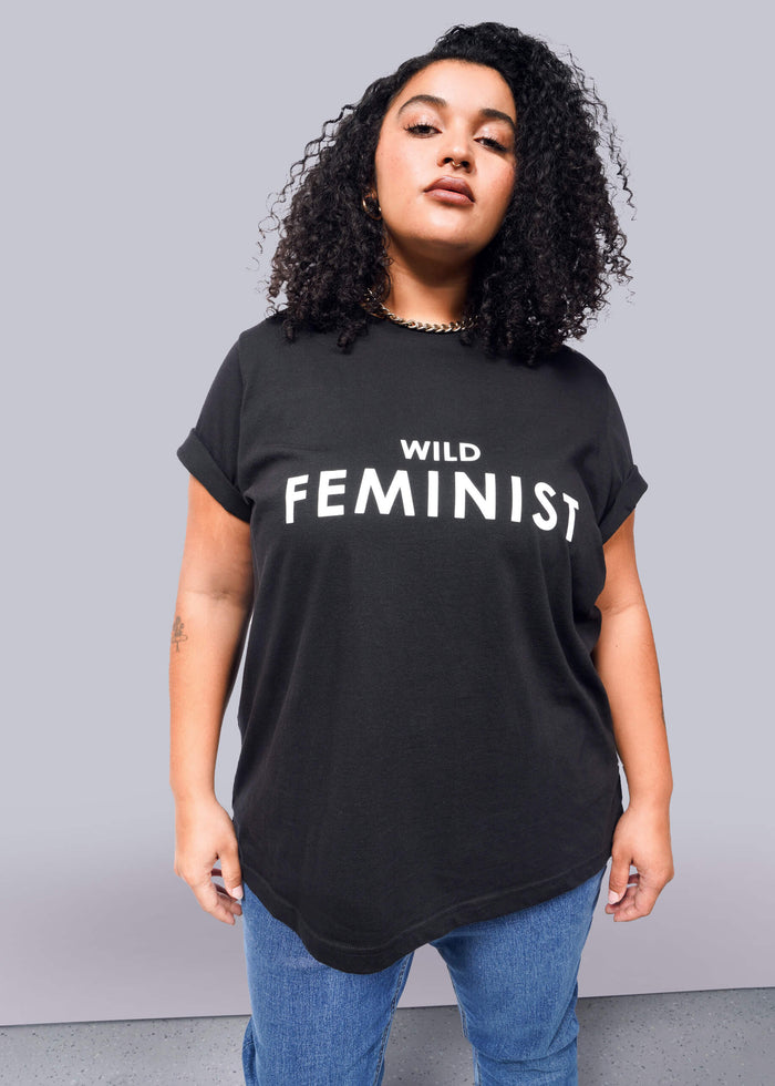 1X=Against a white background, a person with curly hair confidently wears The Wild Feminist® black tee, made of soft cotton and featuring bold white text. Paired with blue jeans, the unisex shirt reflects their expressive style.