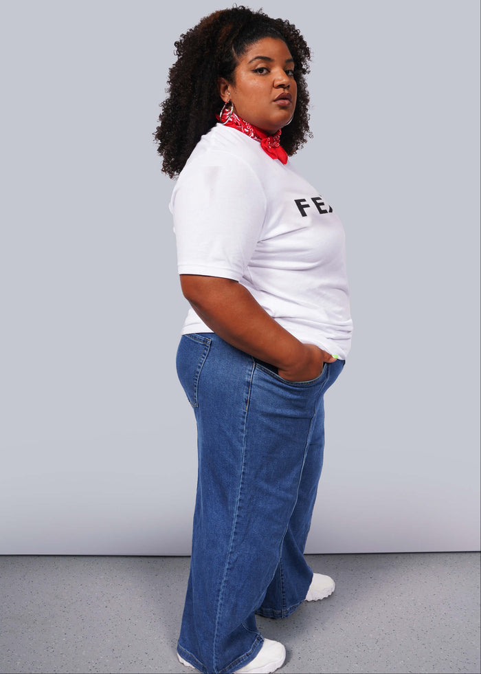 1X=A person with curly hair confidently poses against a gray background, wearing a relaxed unisex fit. They sport The Wild Feminist® Tee, blue jeans, white sneakers, and a red bandana around their neck. One hand in their pocket, they embody unity.