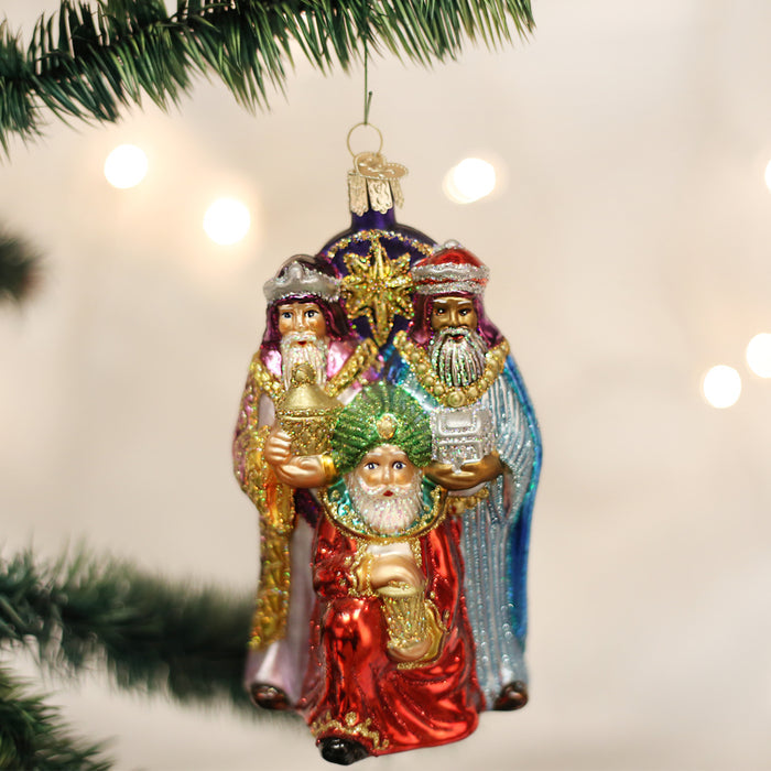 A glass Christmas ornament of the three wise men, dressed in colorful robes and holding gifts, hangs from a green Christmas tree branch with blurred lights in the background.