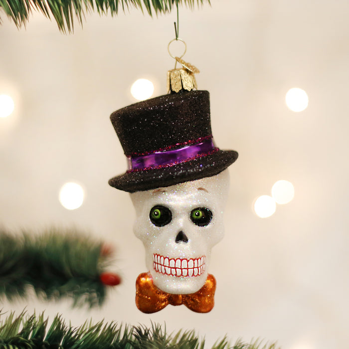 A Christmas tree ornament shaped like a glittery skull with green eyes, a wide toothy grin, a black top hat with a purple band, and an orange bow tie, hanging from a tree branch.