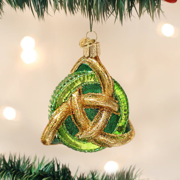 A green and gold glittery Celtic knot ornament hangs from a Christmas tree branch, with blurred white holiday lights in the background.