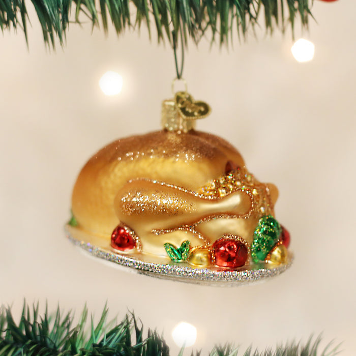A Christmas tree ornament shaped like a roasted turkey on a platter, decorated with glitter and colorful vegetables, hanging from a tree branch.