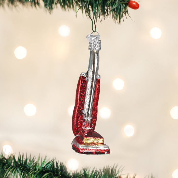 A red and silver vacuum cleaner ornament hangs from a green Christmas tree branch with blurred white holiday lights in the background.