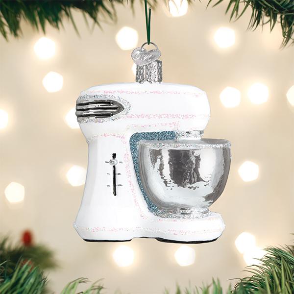 A white stand mixer ornament with a silver mixing bowl hangs on a Christmas tree, with blurred lights in the background and green pine branches framing the image.