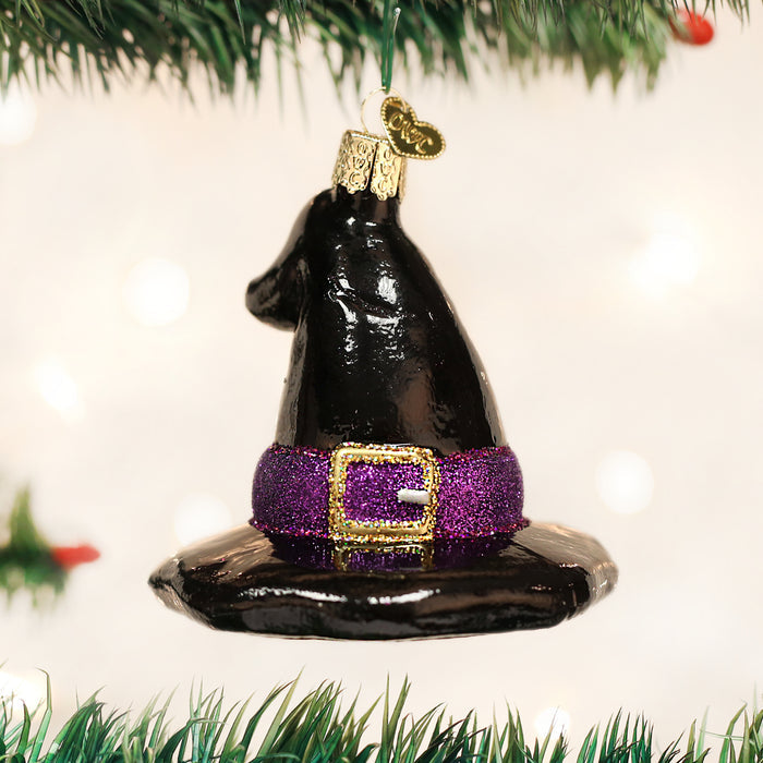 A black witch hat Christmas ornament with a glittery purple belt and gold buckle hangs from a green tree branch against a blurred light background.