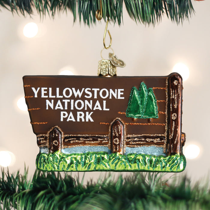 A Christmas tree ornament shaped like a wooden sign with â€œYellowstone National Parkâ€_x009d_ written on it, featuring two green trees, hanging among pine branches with blurred lights in the background.