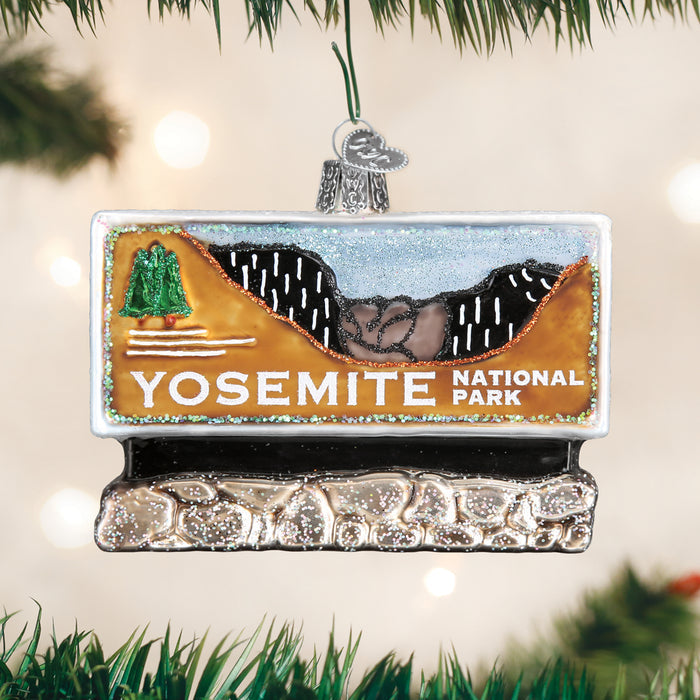 A decorative ornament shaped like a Yosemite National Park sign hangs on a Christmas tree, featuring a scenic mountain landscape, a green tree, and sparkling glitter accents.